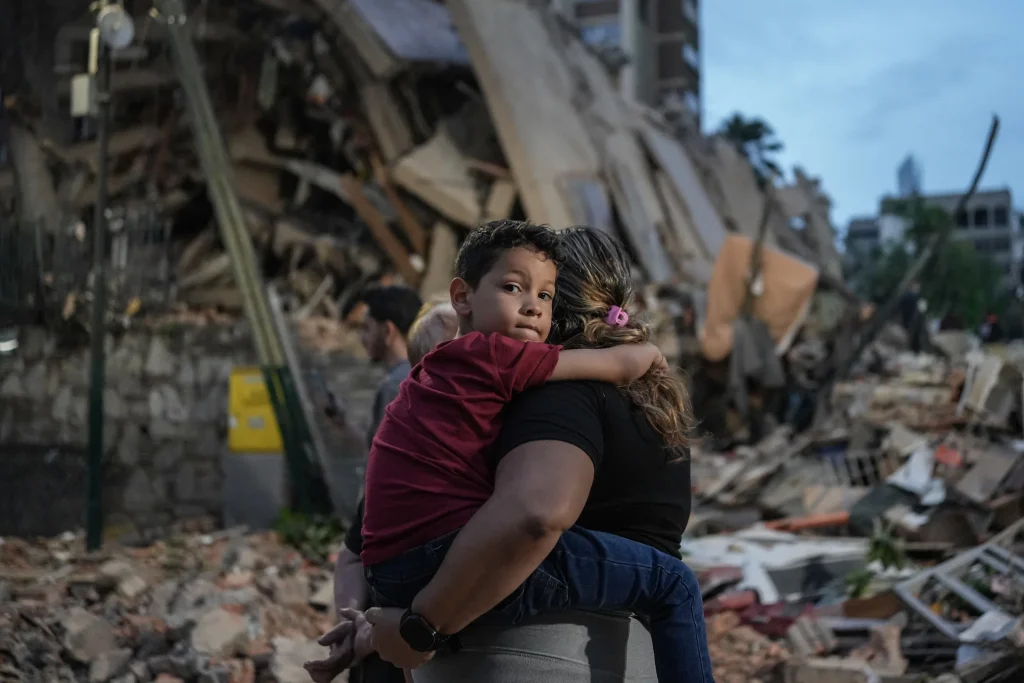 Niño en brazos de su madre frente a los escombros de un edificio residencial colapsado tras el doble terremoto en Venezuela de 2026.